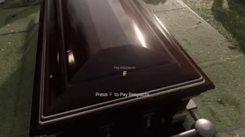 Um caixão num funeral militar, visto de cima, com o aviso do jogo na tela: Press F to Pay Respects. A câmera se aproxima e uma mão de luva branca pousa sobre a tampa.